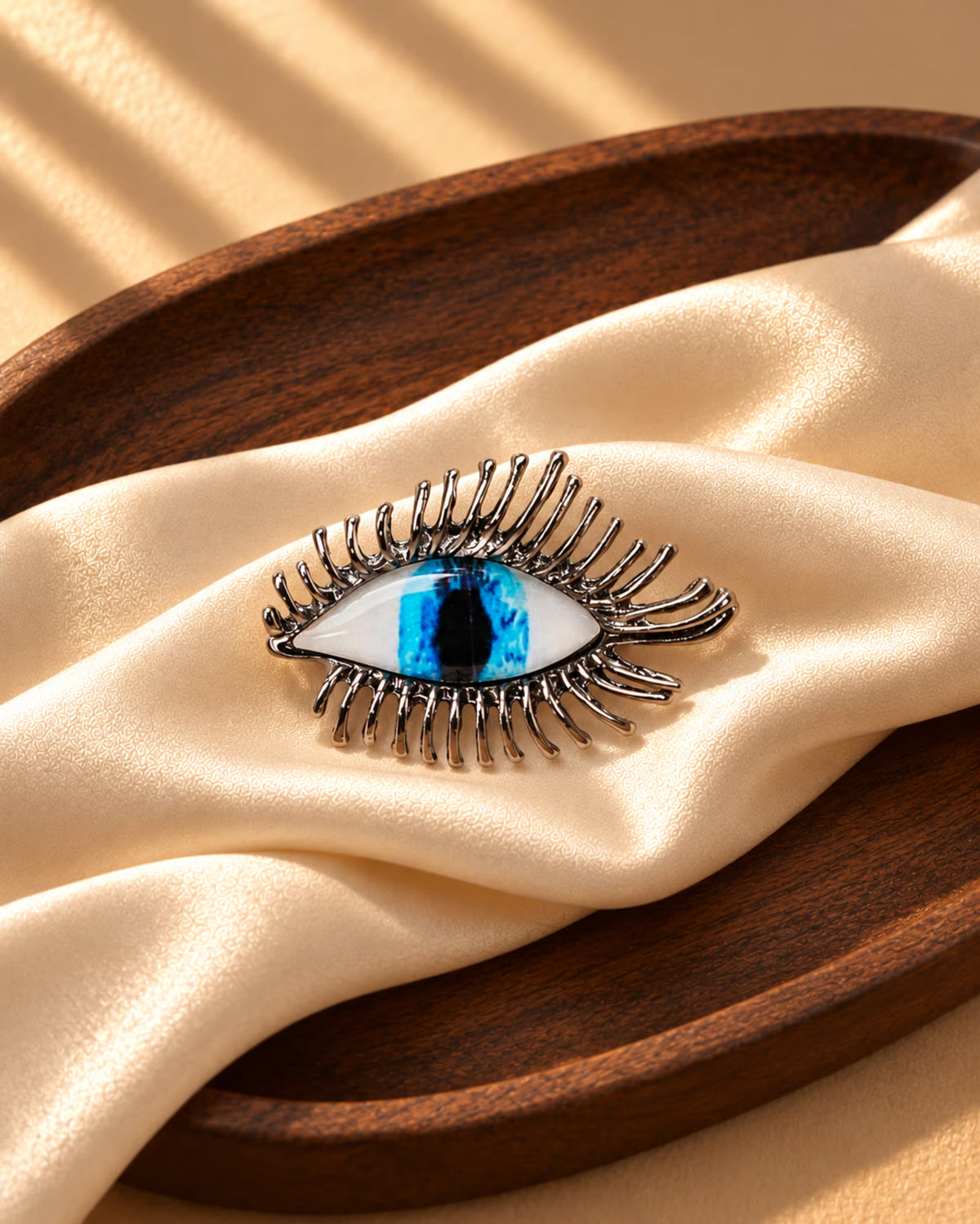 Close-up of Tie Shop Silver Blue Eye Crystal Brooch on beige silk cloth inside a wooden tray, an artistic metallic accessory. صورة مقربة لـ بروش العين الزرقاء الفضي من تاي شوب على قماش حرير بيج وطبق خشبي، اكسسوار معدني فني.
