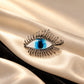 Macro shot of Tie Shop Silver Blue Eye Crystal Brooch with realistic blue iris on beige silk and black velvet background. صورة مقربة جداً لـ بروش العين الزرقاء الفضي من تاي شوب على خلفية ناعمة من الحرير والمخمل.