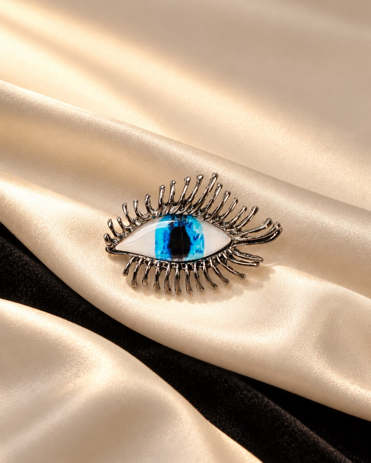 Macro shot of Tie Shop Silver Blue Eye Crystal Brooch with realistic blue iris on beige silk and black velvet background. صورة مقربة جداً لـ بروش العين الزرقاء الفضي من تاي شوب على خلفية ناعمة من الحرير والمخمل.